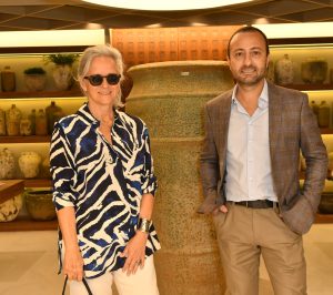 Mulher branca de camisa estampada e cabelos grisalhos ao lado de um vaso alto e homem branco de terno claro